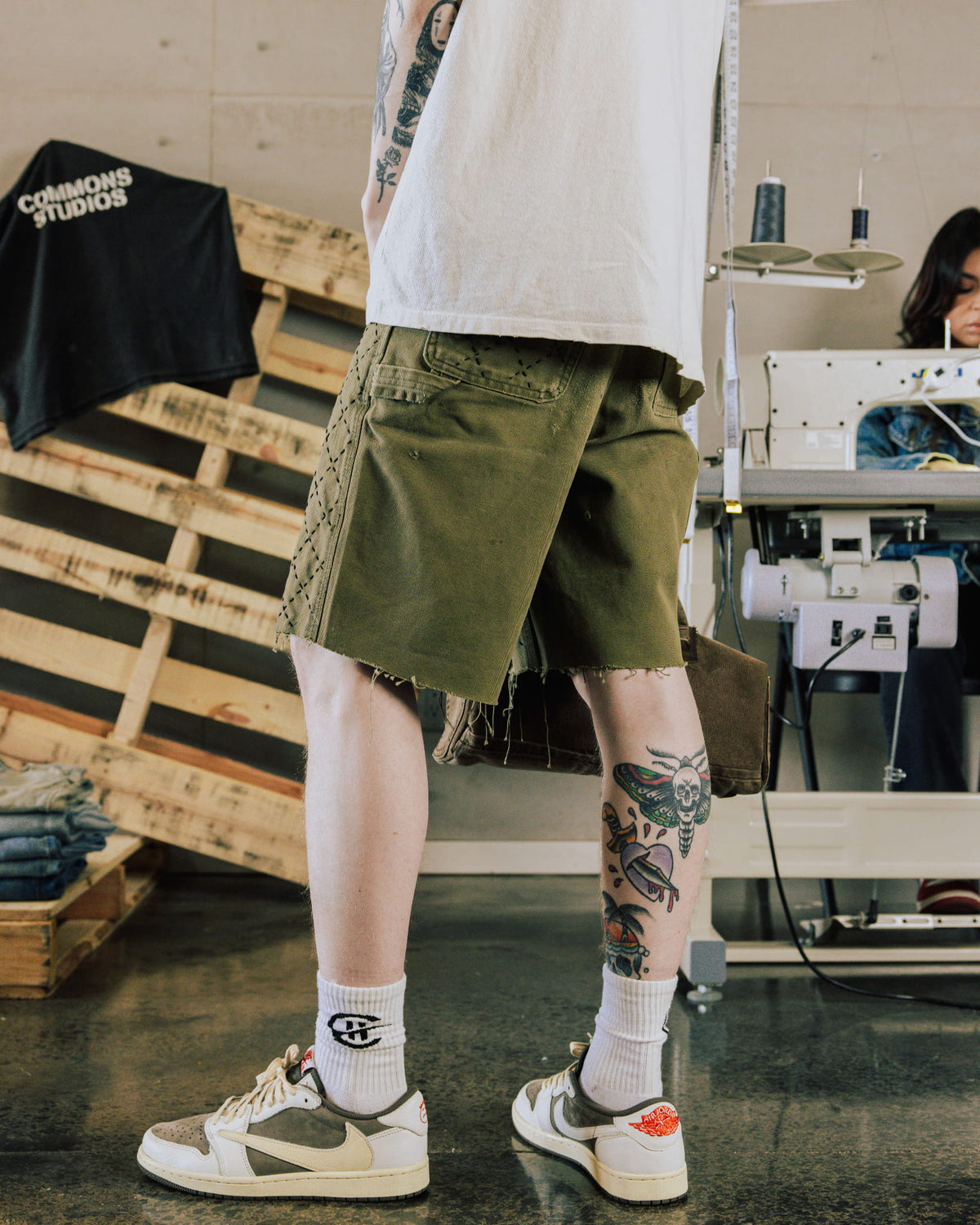 Tattooed person wearing olive green shorts with frayed hems and black stitching beside a sewing machine.
