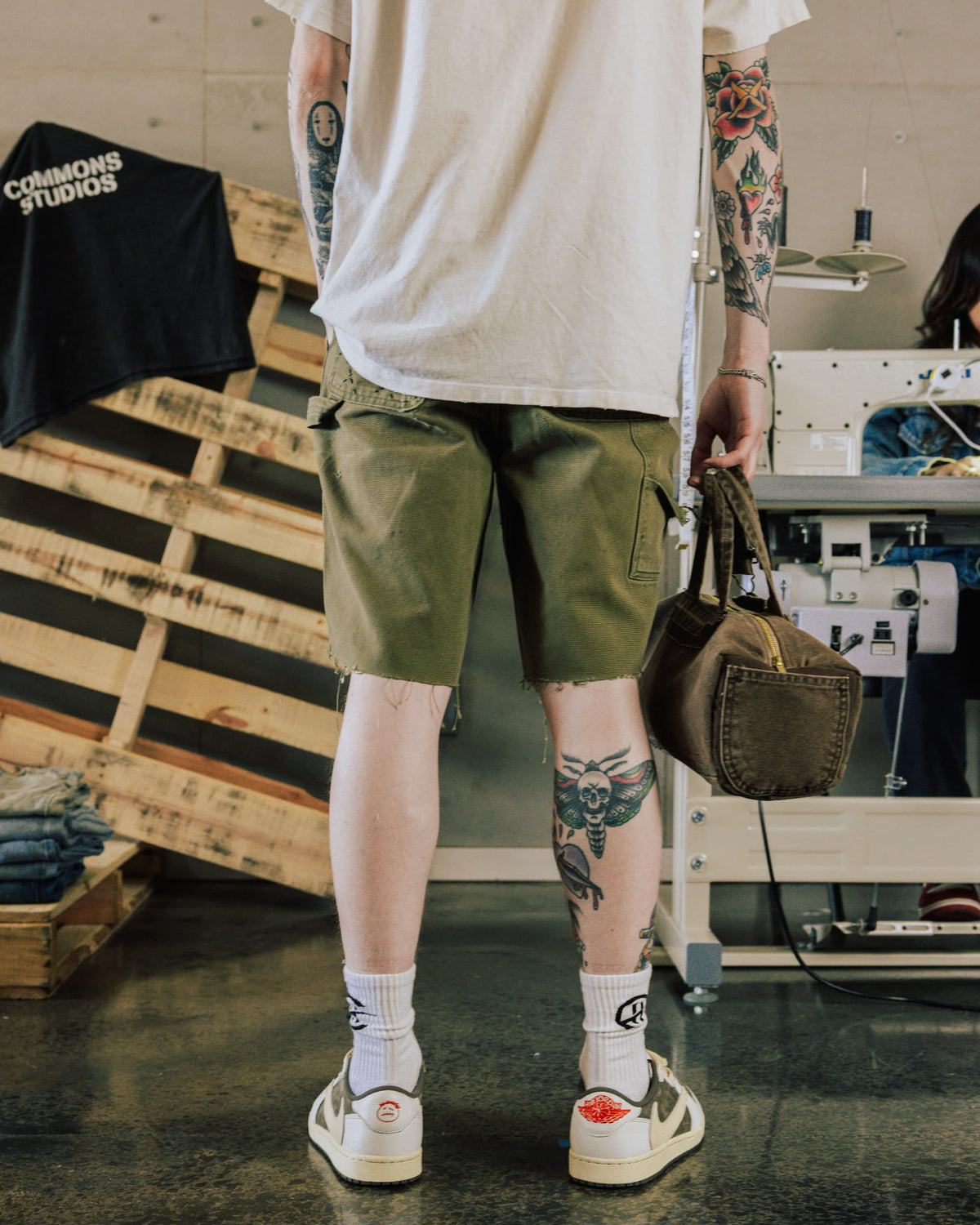 Tattooed person wears a white T-shirt and olive raw-hem shorts, holding a brown zippered bag in a sewing workshop.