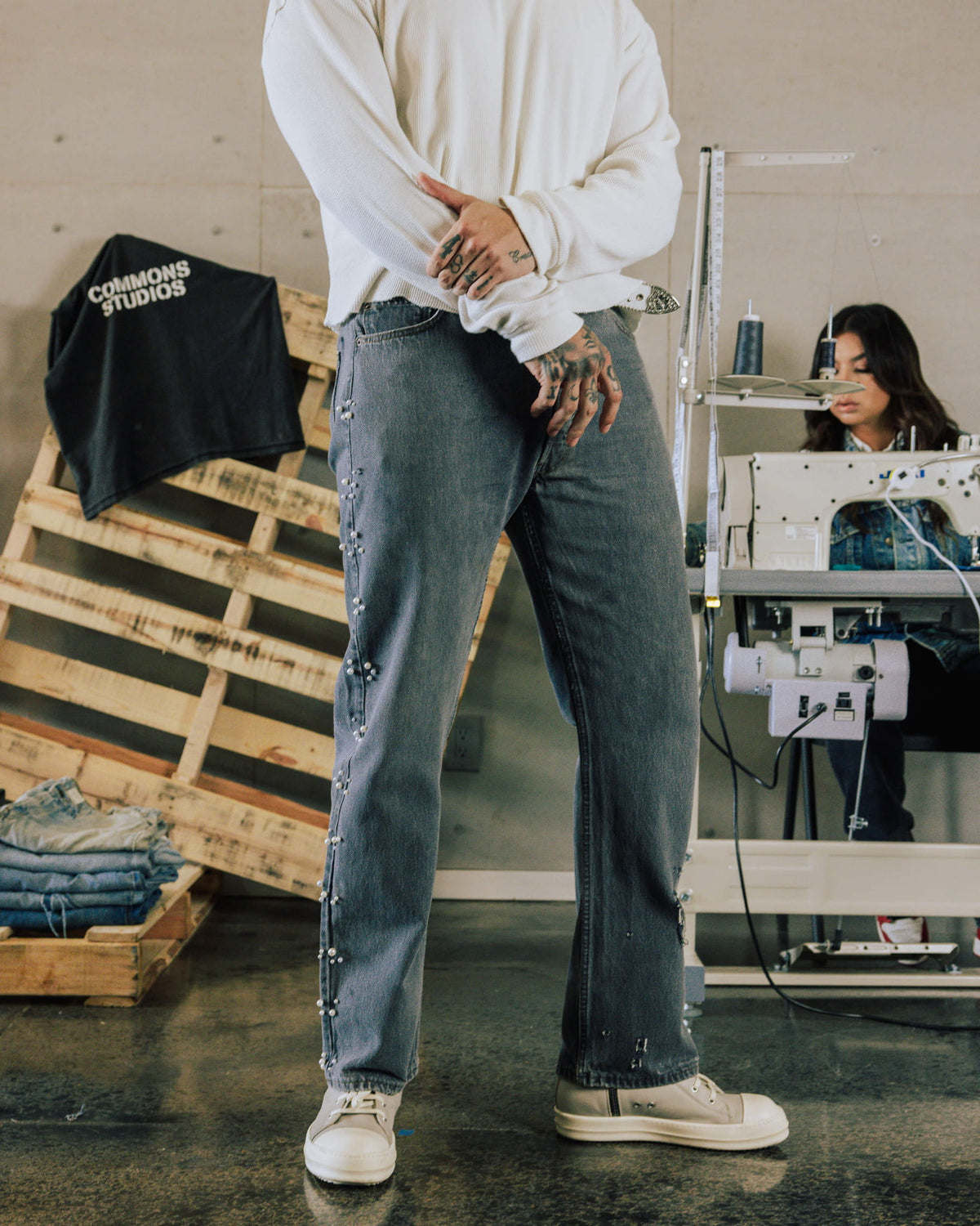 Model wears gray denim jeans with pearl-like side embellishments, a white long-sleeve top and cream shoes in a sewing studio.