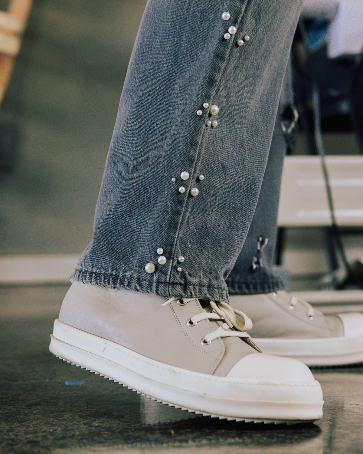Faded blue denim jeans with pearl-like bead clusters along the seams, worn with cream lace-up sneakers on a tiled floor.