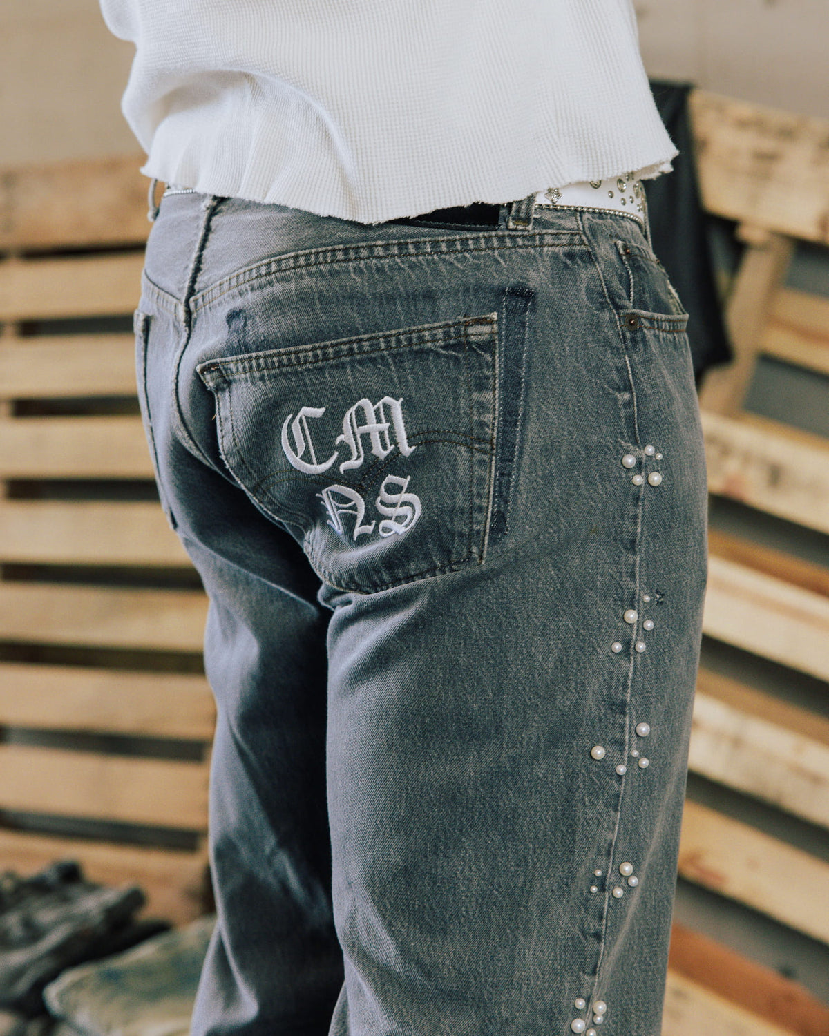 Person wearing faded gray denim jeans with white back-pocket embroidery and pearl-like studs along the side seam.