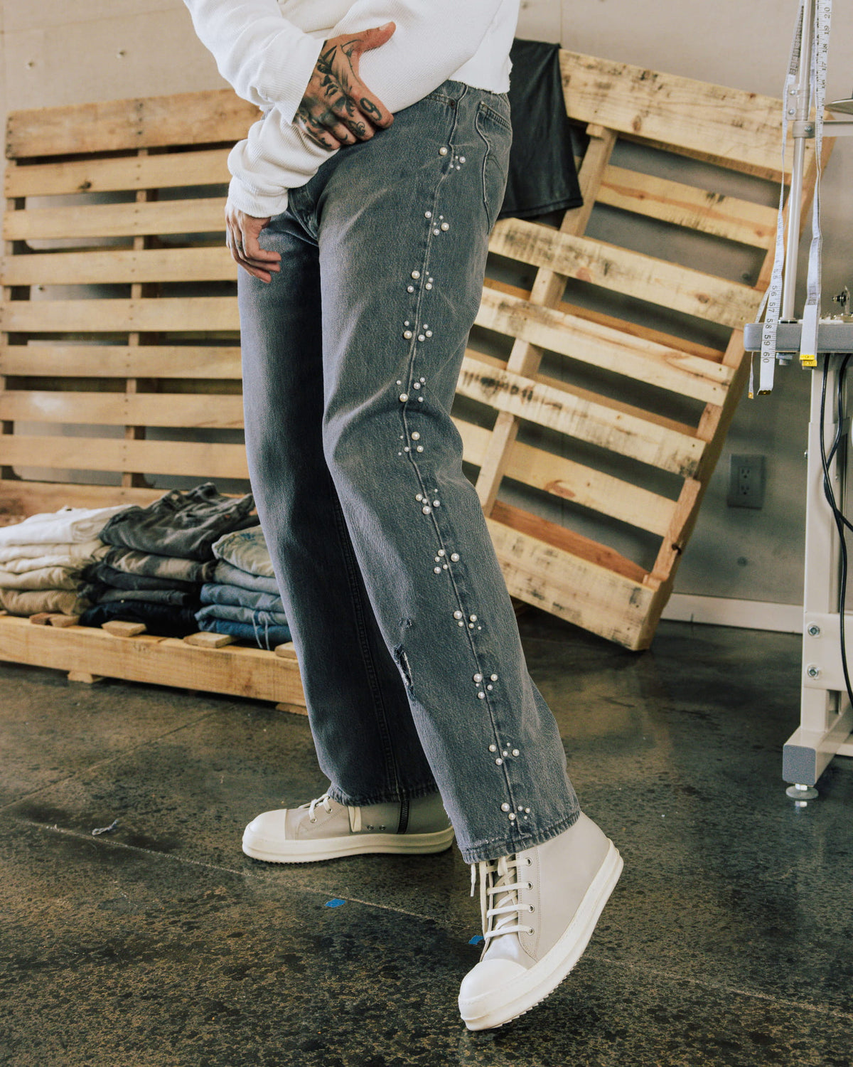Model wears gray denim jeans with pearl-like side embellishments, a white sweater and cream sneakers beside wooden pallets.