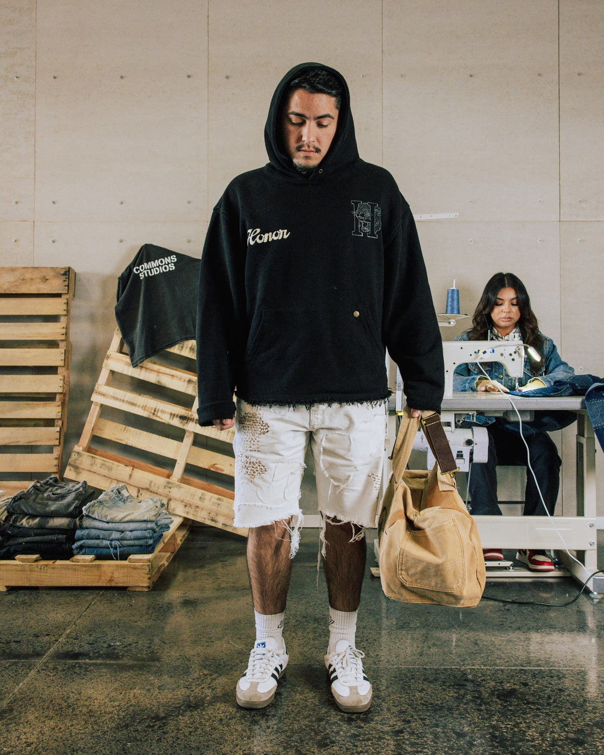 Man wearing a black graphic hoodie and distressed white denim shorts, holding a tan bag in a sewing workshop.