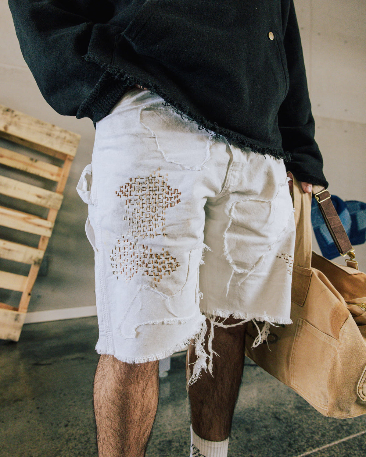 Person wearing white patchwork denim shorts with tan stitching and frayed hems, a black top, and carrying a tan canvas bag.