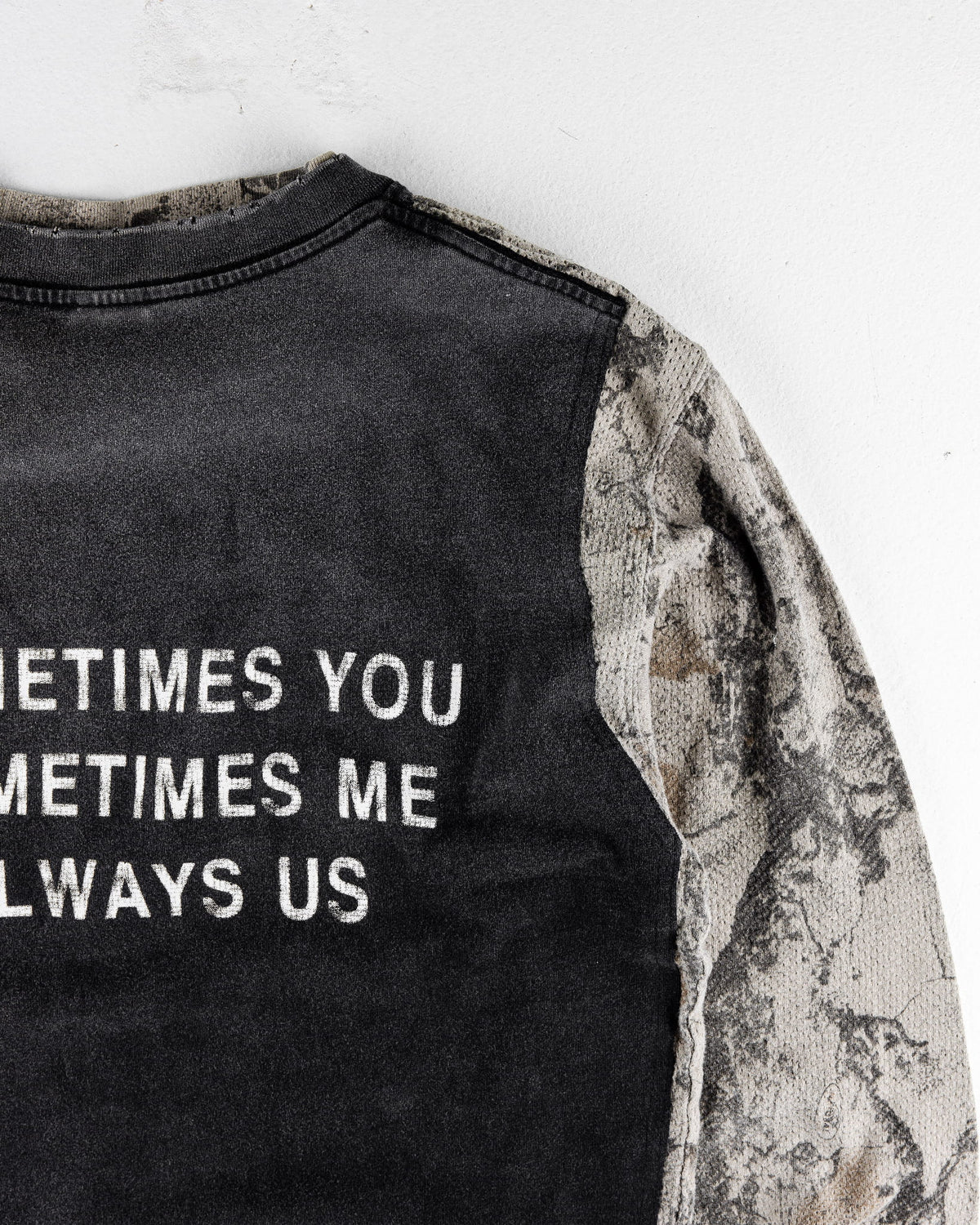 Close-up of a black knit shirt with white lettering and gray camouflage sleeves on a white surface.