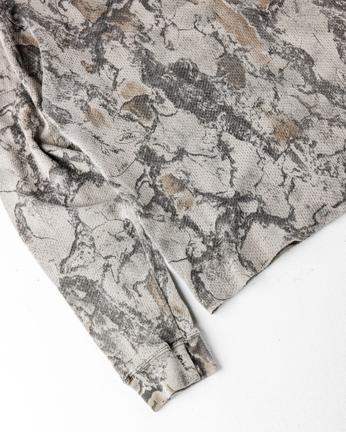 Close-up of a beige, gray and brown camouflage waffle-knit long-sleeve shirt with a cuff on a white surface.