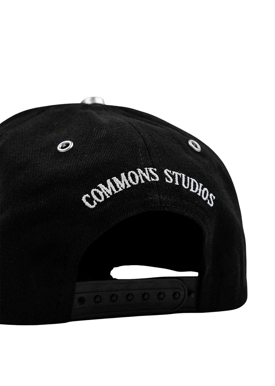 Back of a black snapback cap with white COMMONS STUDIOS embroidery, silver eyelets, and an adjustable plastic strap.