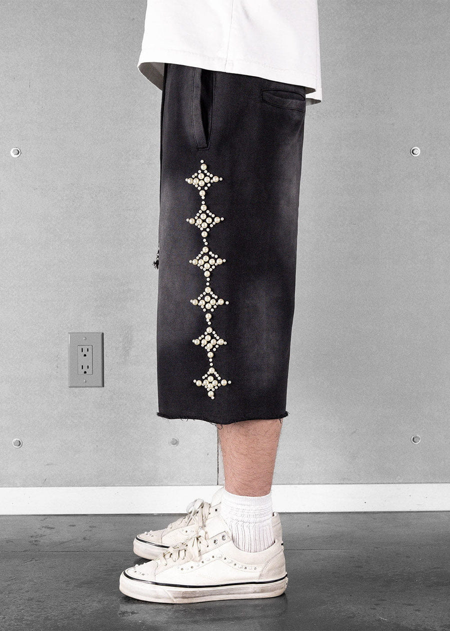 Black long shorts with pearl-like diamond embellishments and raw hems, worn with a white shirt and white sneakers.