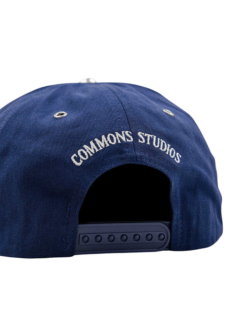 Back of a navy blue snapback cap with white COMMONS STUDIOS embroidery, metal eyelets, and an adjustable plastic closure.