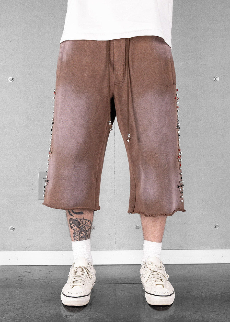 Brown wide-leg drawstring shorts with raw hems and beaded side details, worn with a white shirt and cream sneakers.