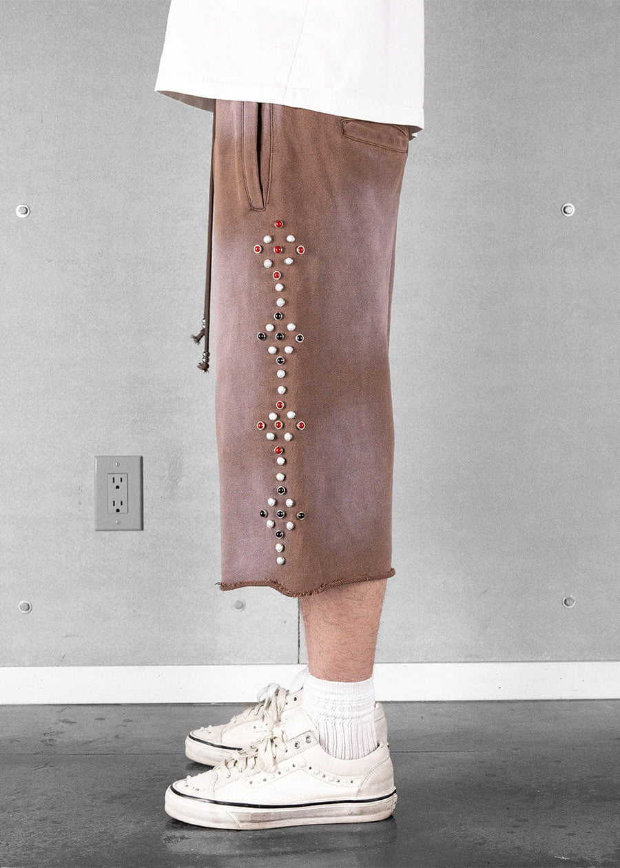 Model wears brown long shorts with red, white and black stud patterns, raw hems and white sneakers against a gray wall.