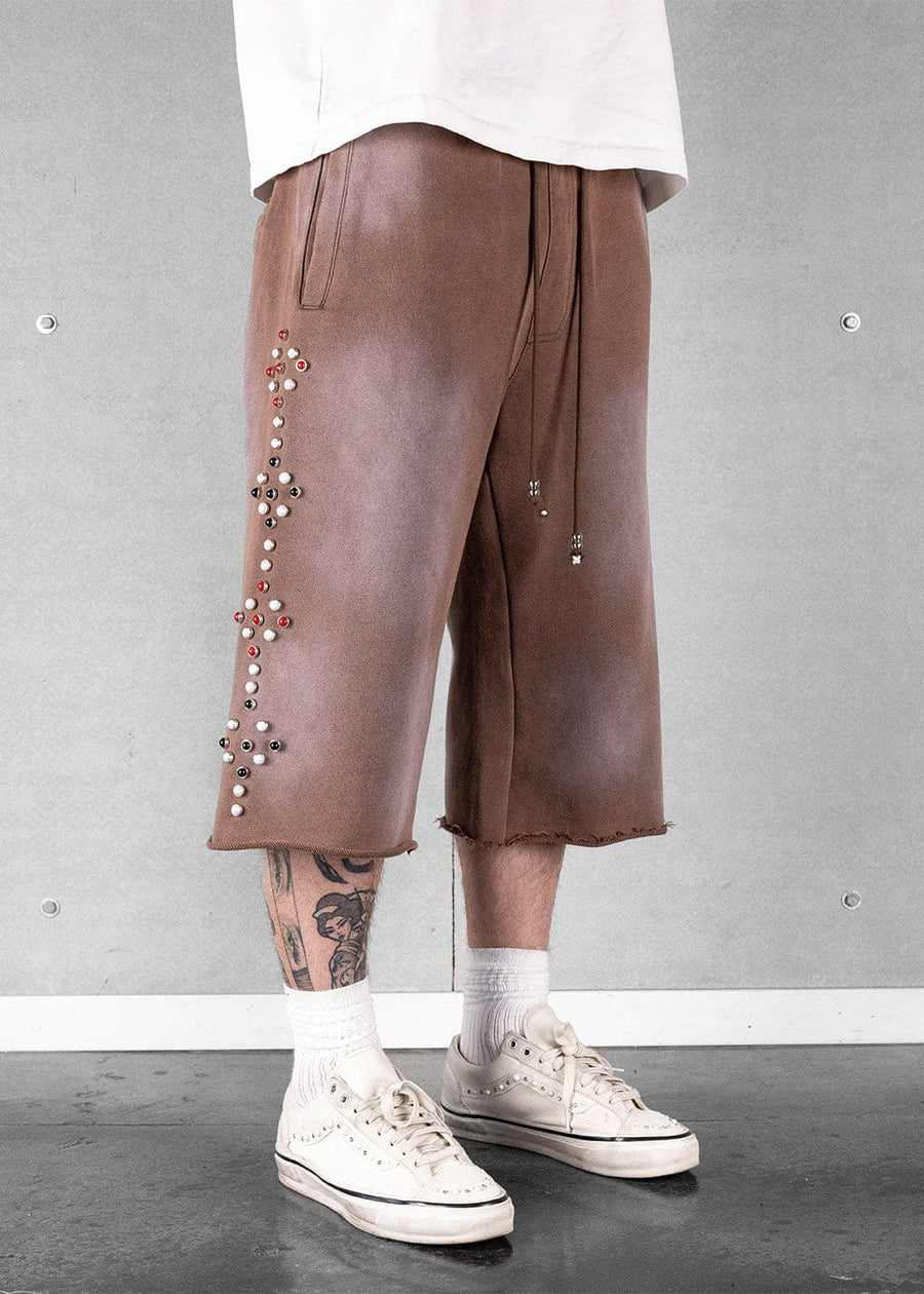 Model wears brown wide-leg drawstring shorts with raw hems and multicolored studs, a white shirt, and white sneakers.