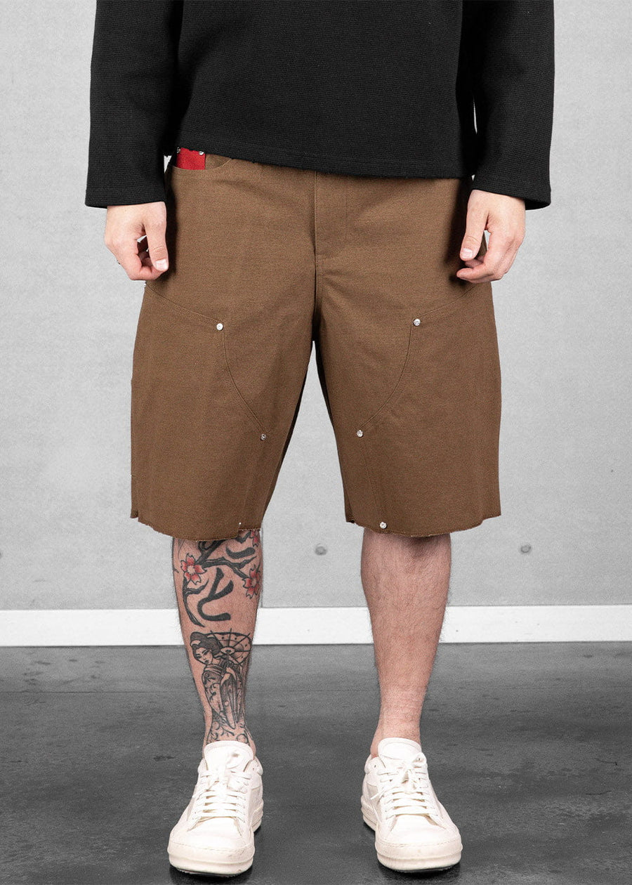 Model wears brown knee-length shorts with riveted front panels and raw hems, a black top, and white sneakers.