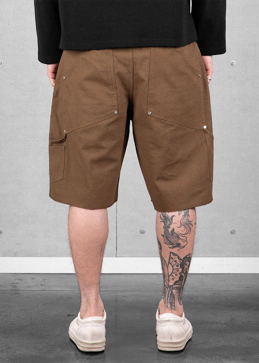 Back view of brown knee-length carpenter shorts with riveted pockets and a hammer loop, worn with a black top and white shoes.