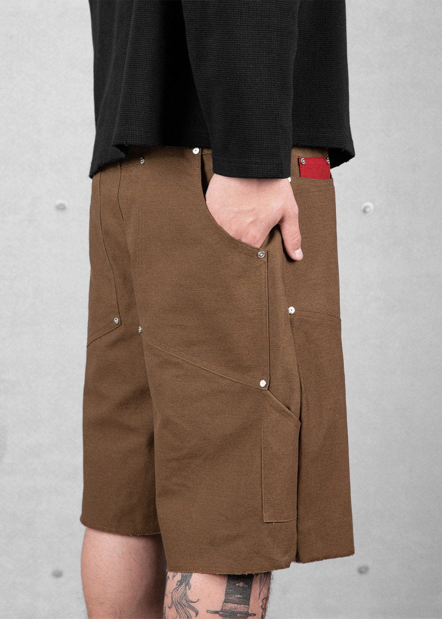 Brown canvas work shorts with riveted pockets and a side loop, worn with a black top against a gray wall.