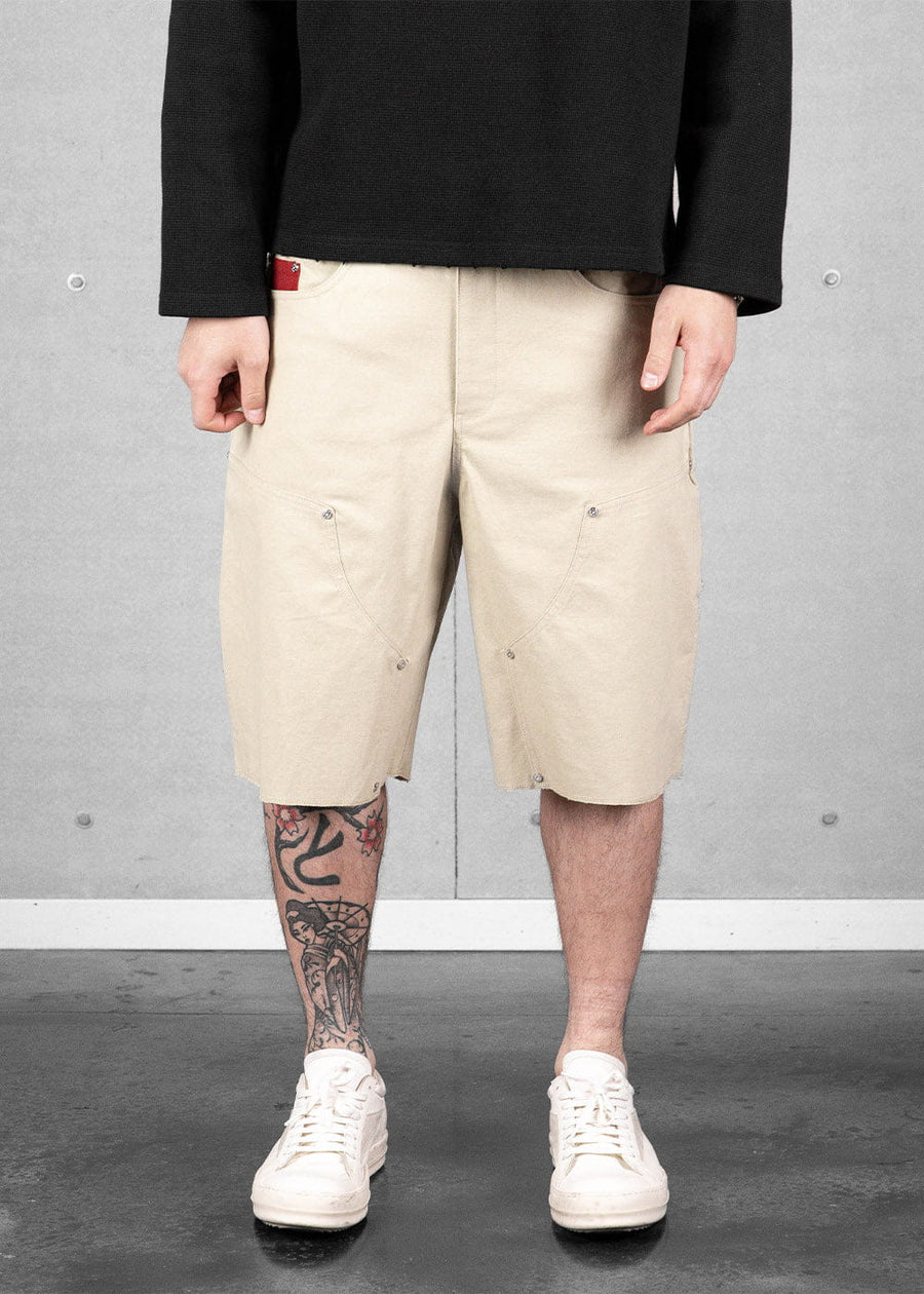 Model wears cream canvas knee-length shorts with curved seams and metal rivets, a black top, and white sneakers.