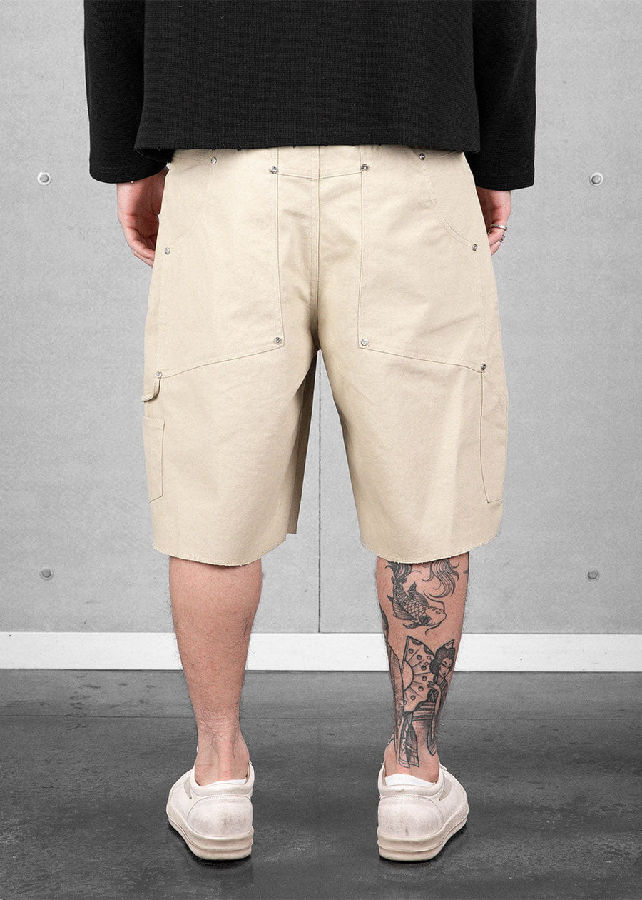 Rear view of cream canvas knee-length shorts with riveted pockets and a side loop, worn with a black top and white sneakers.