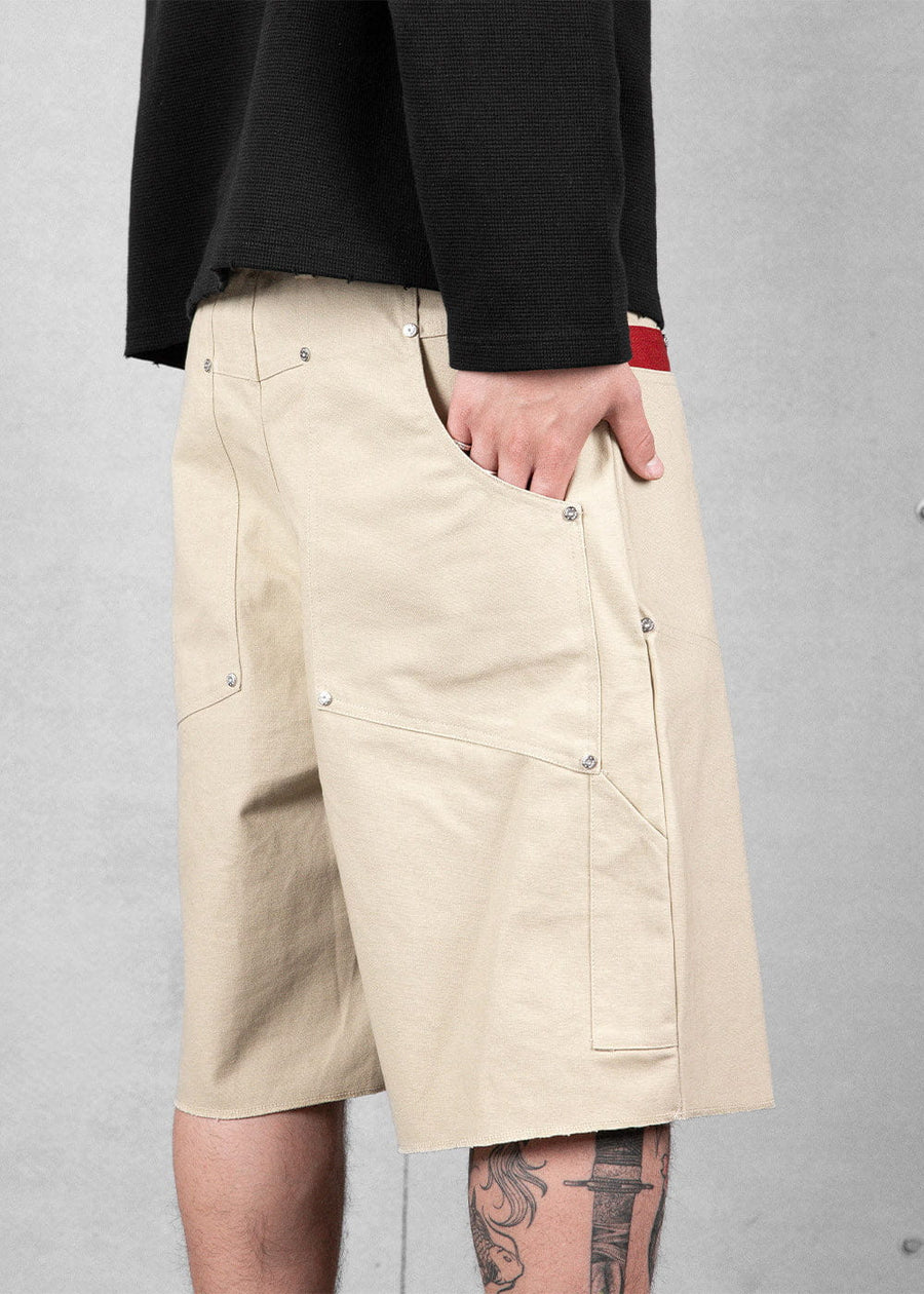 Cream canvas knee-length shorts with riveted patch pockets and raw hems, worn with a black top against a gray wall.