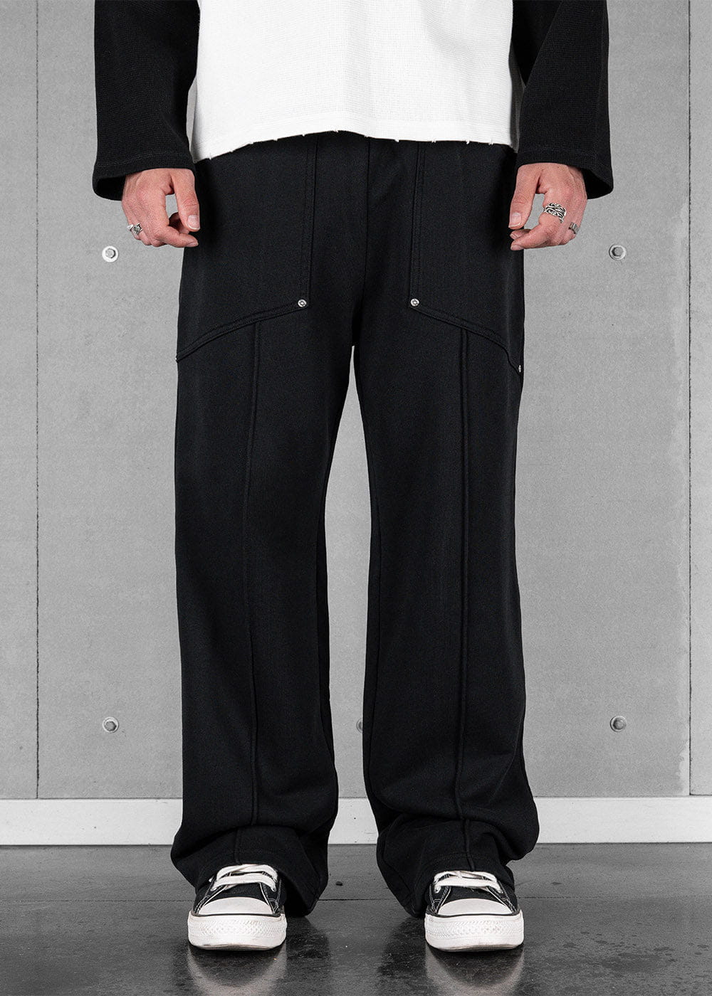 Model wearing black wide-leg sweatpants with front seams and silver rivets, paired with black-and-white sneakers.