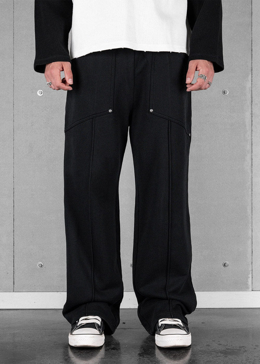 Model wearing black wide-leg sweatpants with front seams and silver rivets, paired with black-and-white sneakers.