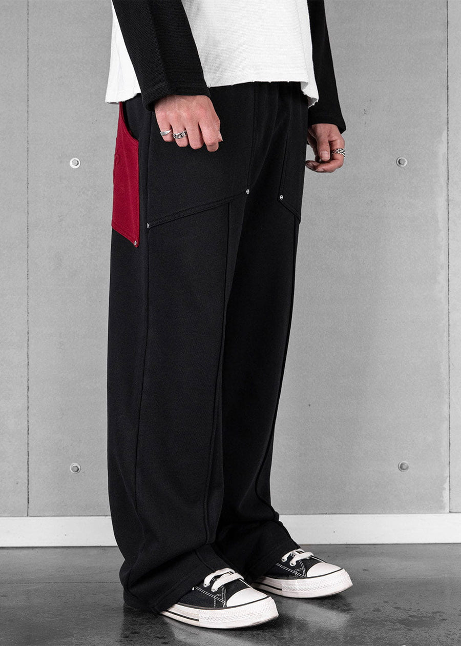 Model wears wide-leg black sweatpants with a red side panel and seam detailing, plus black sneakers against a gray wall.