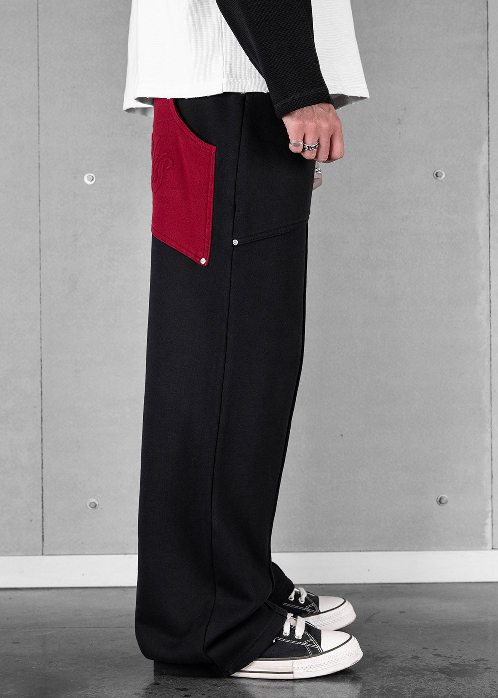 Model wears black wide-leg sweatpants with a large red patch pocket and silver rivets against a gray concrete wall.