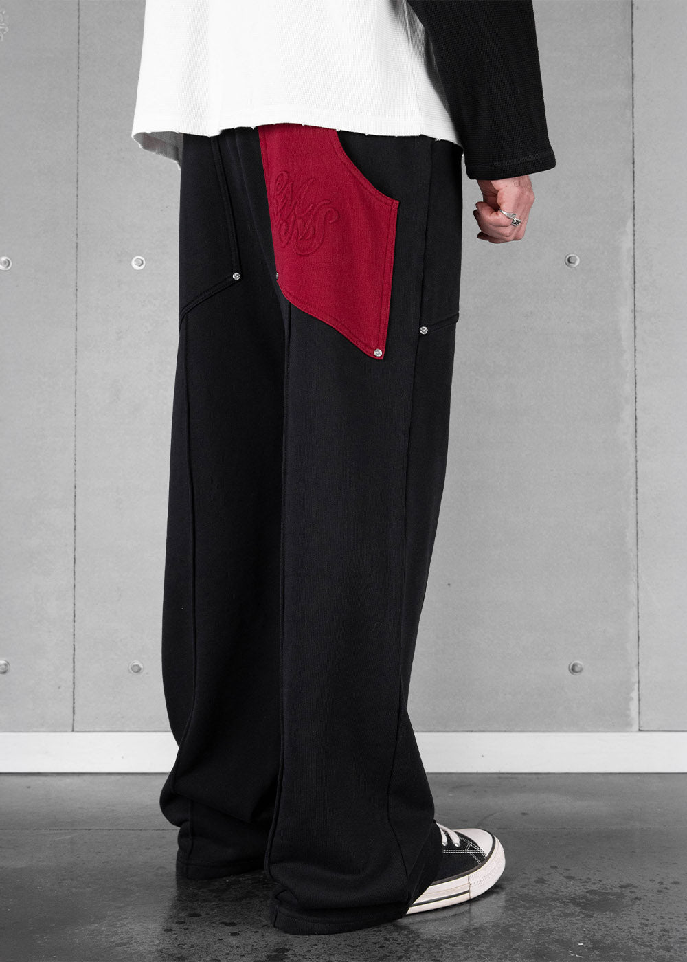 Model wearing wide-leg black sweatpants with a large red embroidered pocket and silver rivets against a gray wall.