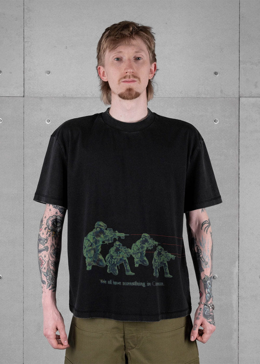 Model wearing a black crewneck T-shirt with green soldier graphics and olive trousers against a gray concrete wall.