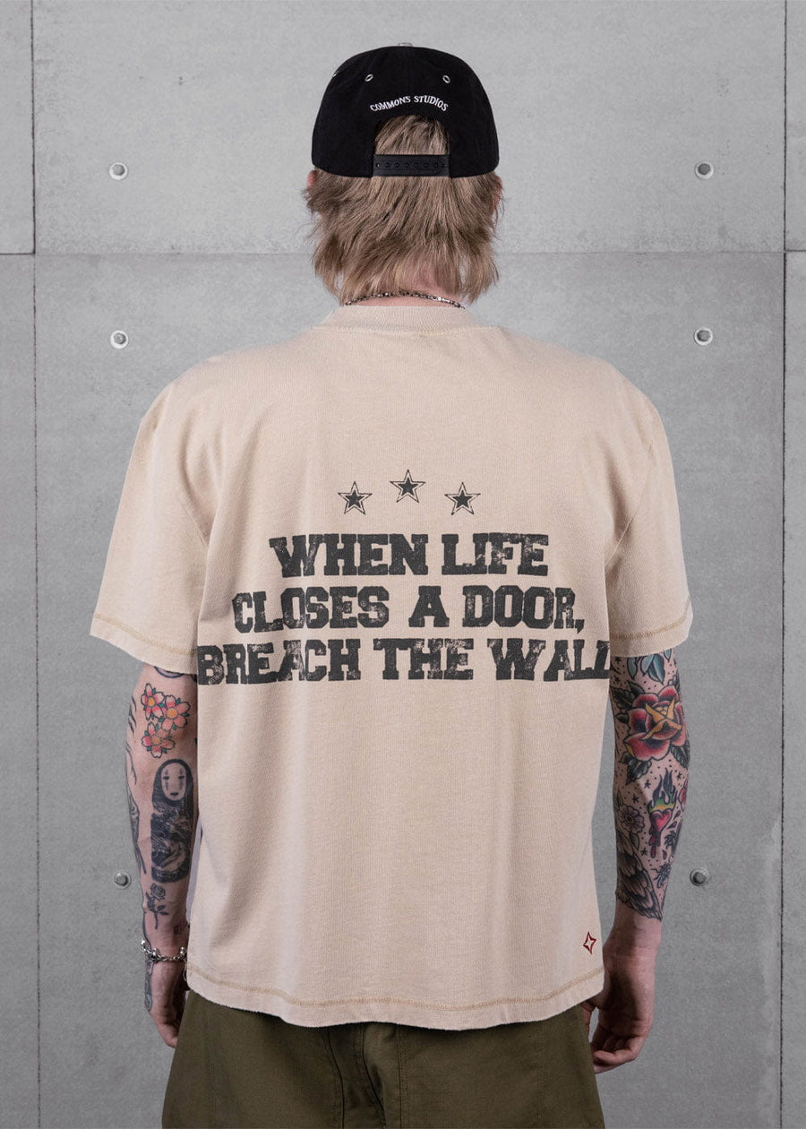 Back view of a person wearing a beige T-shirt with black slogan and three stars, a black cap, and olive trousers.