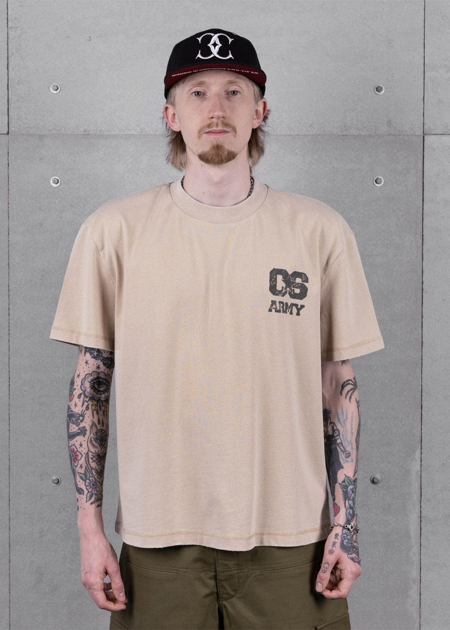 Model wearing a beige crewneck T-shirt with a dark chest graphic, olive pants and a black cap against a gray concrete wall.