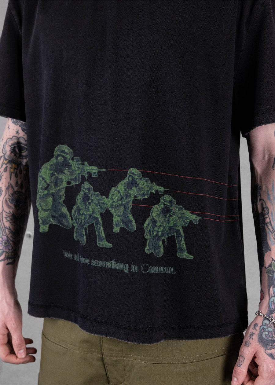 Black T-shirt with green soldier graphics, thin red lines and text, worn by a tattooed model with olive pants.