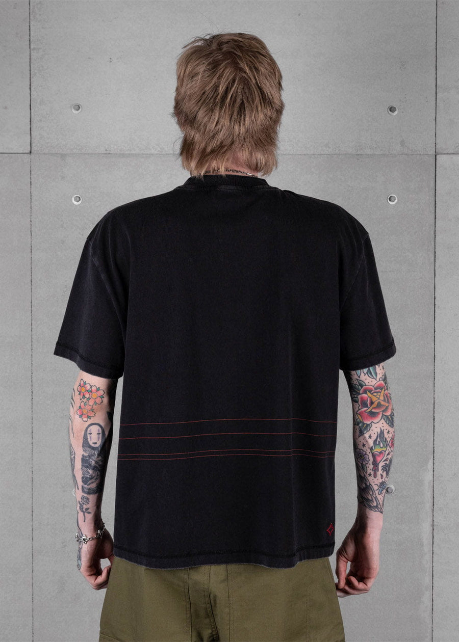 Back view of a tattooed model wearing a black T-shirt with thin red lines and olive pants against a gray concrete wall.