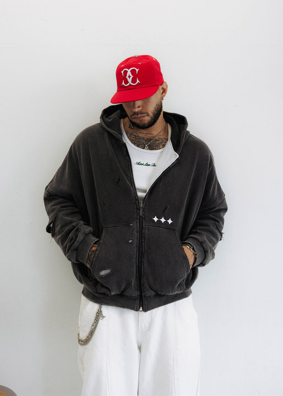 Man wearing a distressed black zip-up hoodie with white star details, a red cap and white pants against a white wall.