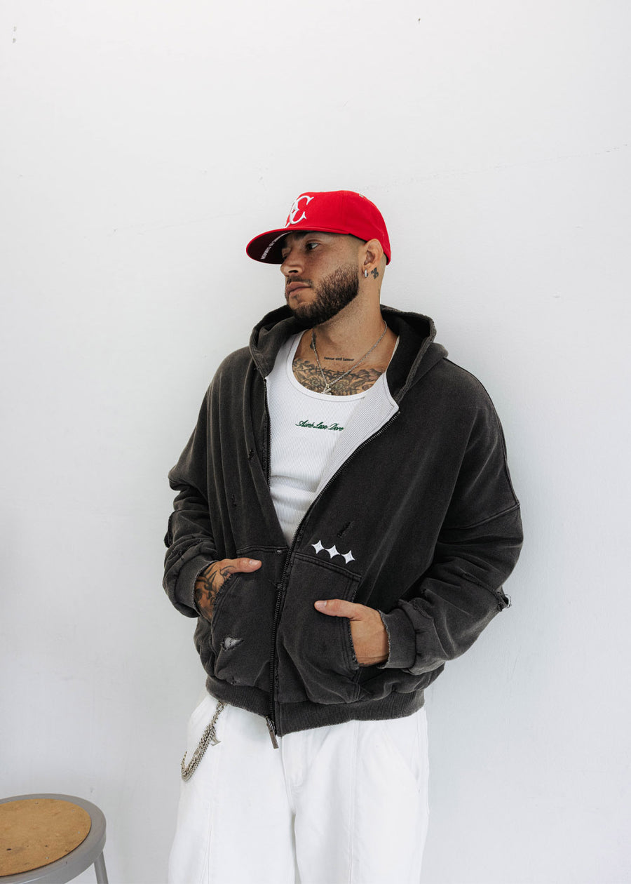 Man wearing a distressed black zip-up hoodie with white star details, white pants and a red cap against a white wall.