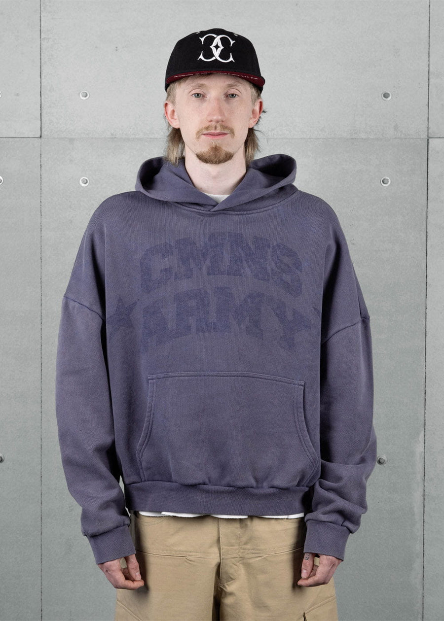 Model wearing a muted blue hoodie with tonal lettering and a kangaroo pocket, black cap and tan pants against a gray wall.