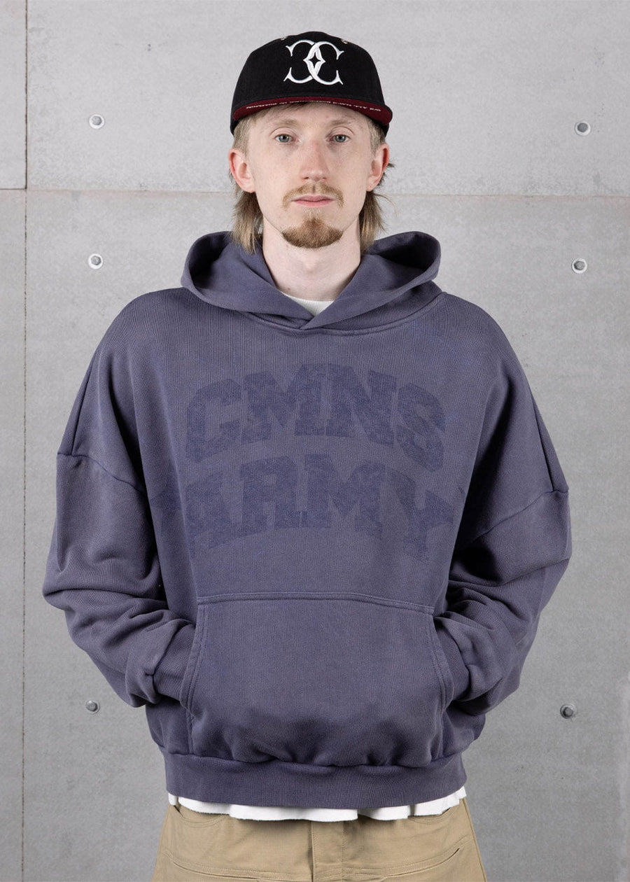 Person wearing a muted blue hoodie with tonal lettering and a kangaroo pocket, plus a black cap, against a concrete wall.