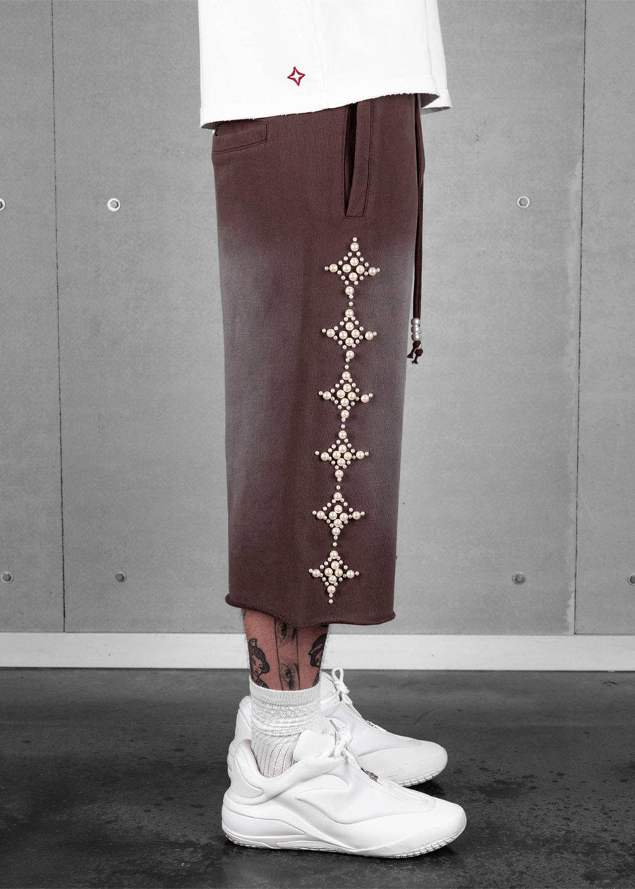 Model wearing brown cropped knit pants with pearl-like star motifs, a white shirt and white sneakers against a gray wall.