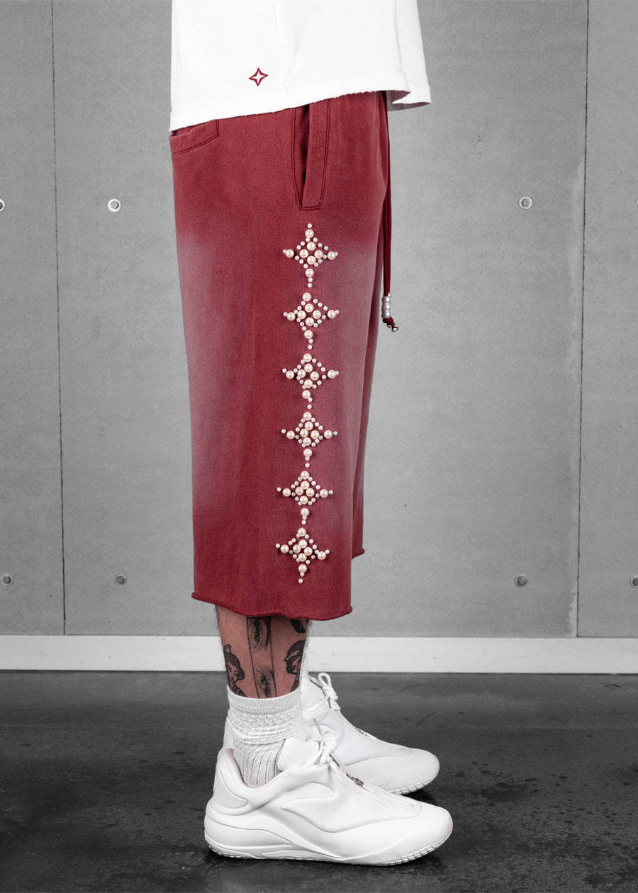 Model wears burgundy long shorts with pearl-like star embellishments, a white T-shirt and white sneakers against a gray wall.