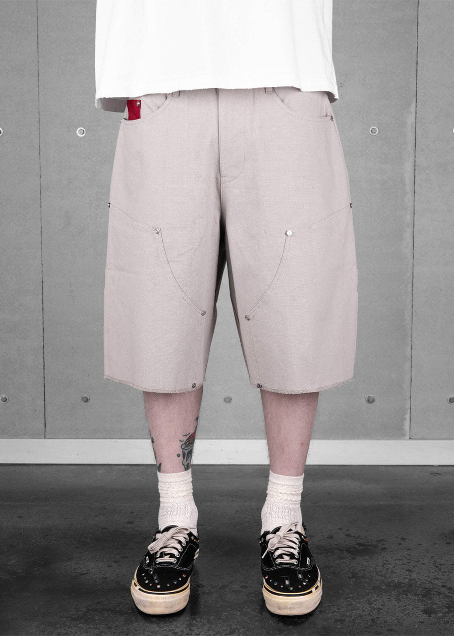 Person wearing light gray canvas knee-length shorts with curved seams and metal rivets against a concrete wall.
