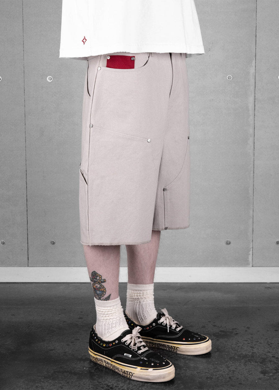 Model wearing gray canvas knee-length shorts with metal rivets, curved seams, and a red pocket accent against a concrete wall.