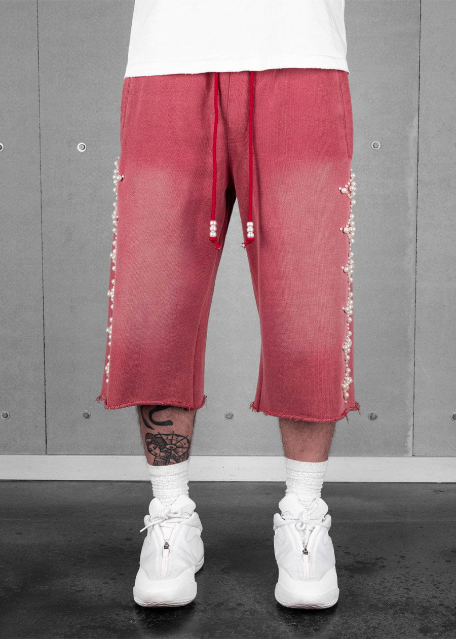 Model wearing faded red long shorts with pearl bead trim, drawstrings and raw hems, paired with white sneakers.
