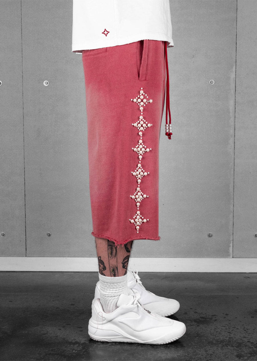 Model wearing faded red long shorts with pearl star motifs, raw hems and beaded drawstrings, paired with white sneakers.