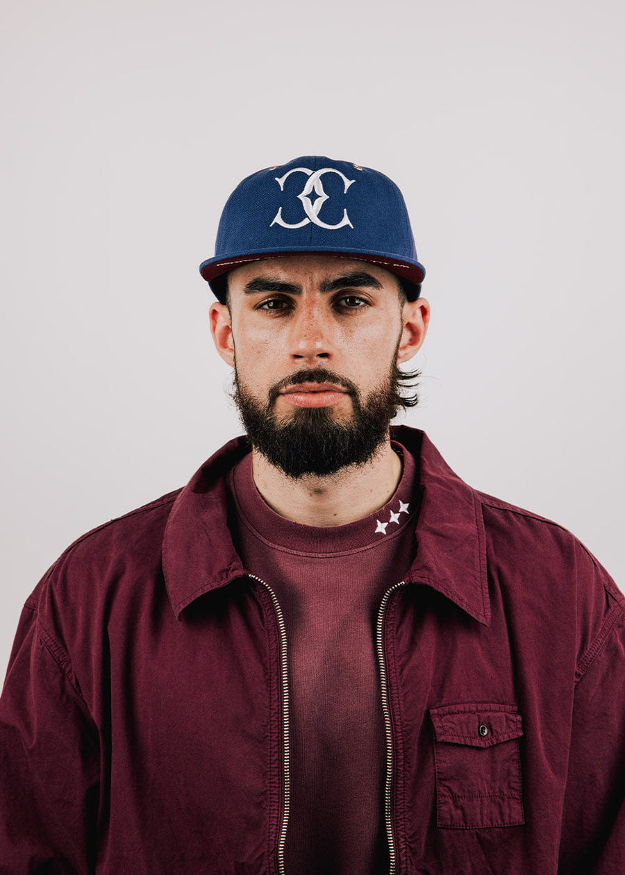 Man wearing a navy flat-brim cap with a white embroidered logo and burgundy clothing against a light gray background.