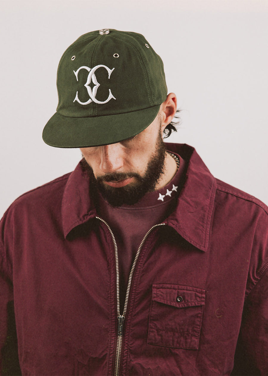 Man wearing an olive green cap with white embroidered logo and metal eyelets, paired with a burgundy zip jacket.