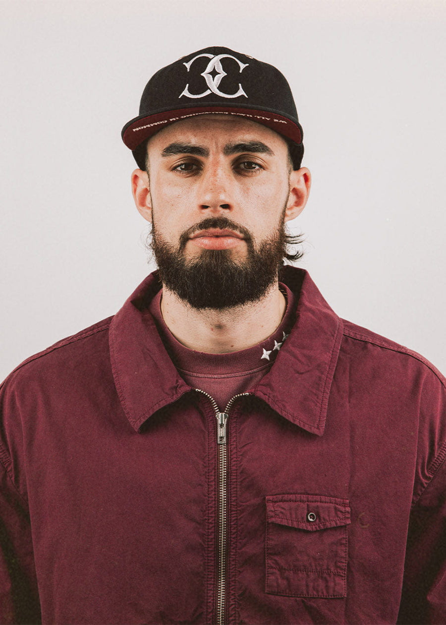 Man wearing a black cap with a white embroidered logo and burgundy underbrim, paired with a burgundy zip-front jacket.