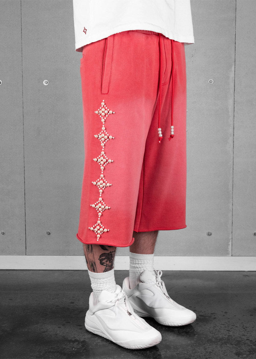 Coral knit long shorts with pearl star motifs and beaded drawstrings, worn with white sneakers against a gray wall.