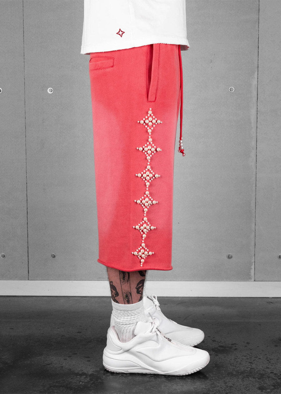 Coral red long shorts with pearl-like star embellishments and drawstrings, worn with a white shirt and sneakers.