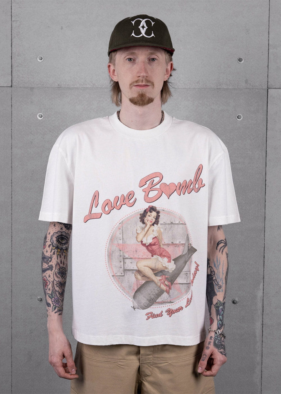 Model wearing a white Love Bomb pinup graphic T-shirt, black cap and tan pants against a gray concrete wall.