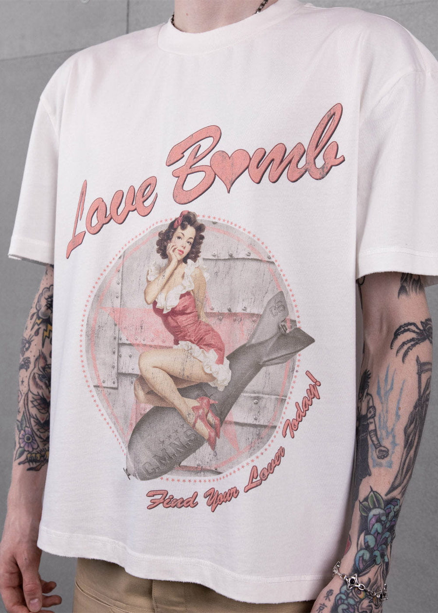 Tattooed model wearing a white crewneck T-shirt with pink Love Bomb lettering and a vintage pinup graphic.