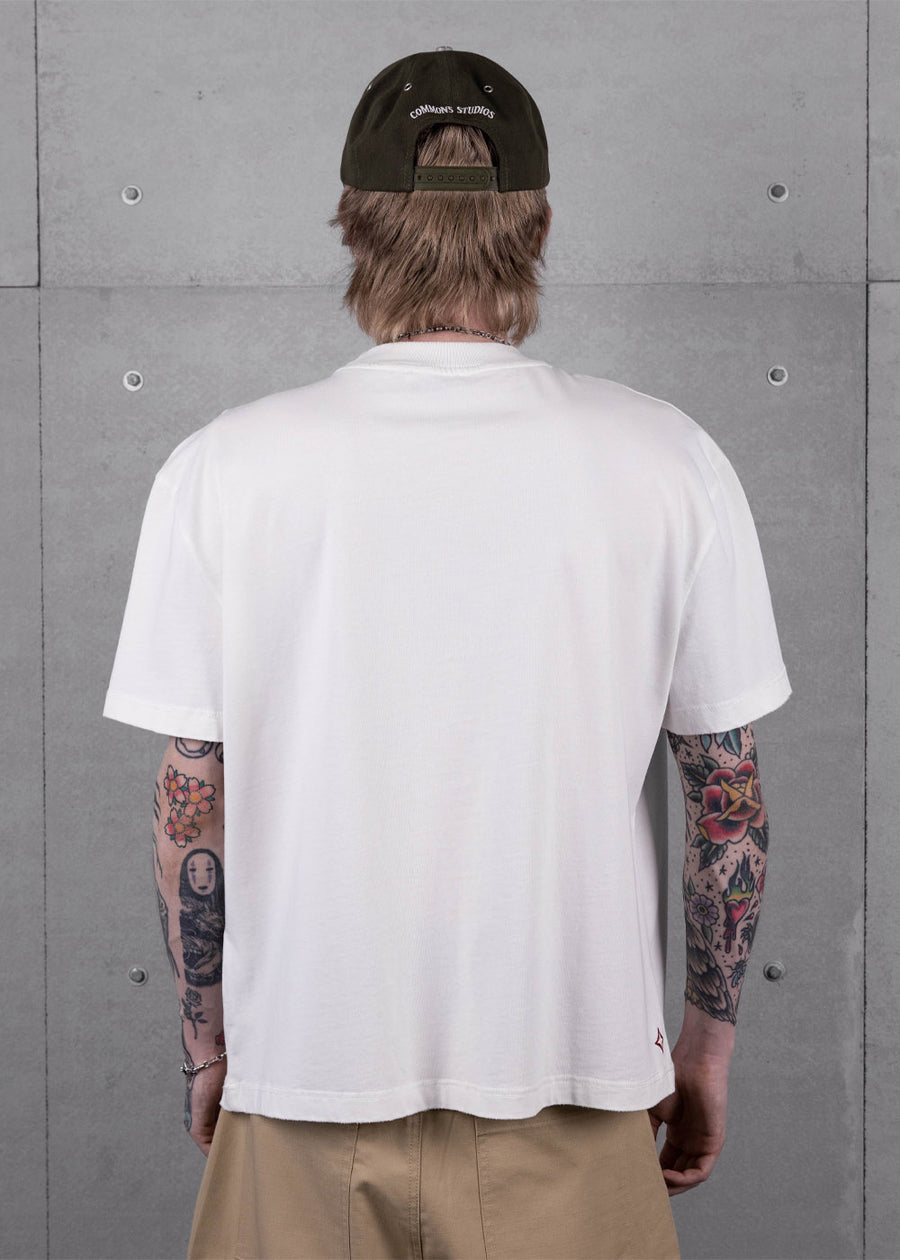 Back view of a tattooed person wearing a loose white T-shirt, tan pants and dark snapback cap against a gray concrete wall.